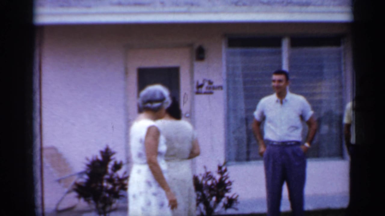 1958 Florida frame with two women in light dresses facing a man in a short-sleeve shirt outside a pink stucco residence.