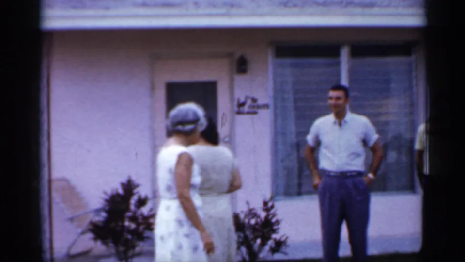 1958 Florida frame with two women in light dresses facing a man in a short-sleeve shirt outside a pink stucco residence.