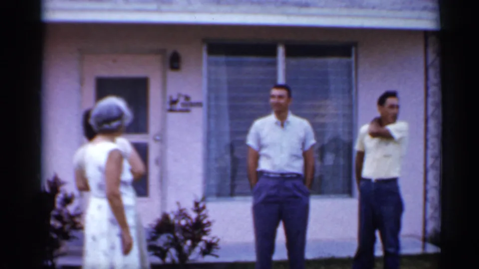 1958 Florida home-movie frame showing two men in short-sleeve shirts and a woman near a pink stucco house exterior.