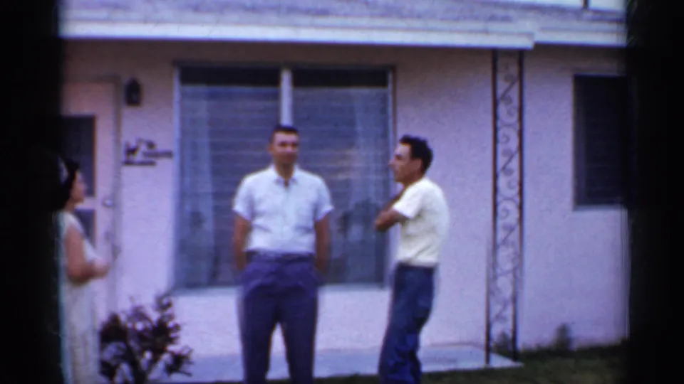 1958 Florida frame showing two men conversing outside a pink stucco home, with a woman partly visible near the doorway.