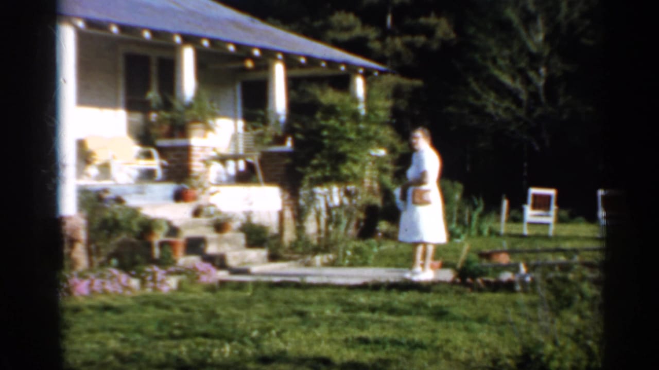 Woman in a white dress standing on a lawn near a small house and garden.