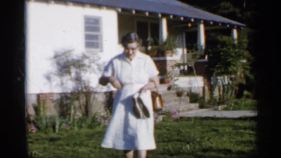 Close 1958 Florida home-movie view of a woman in a white dress holding a purse or shoes near a house and garden.