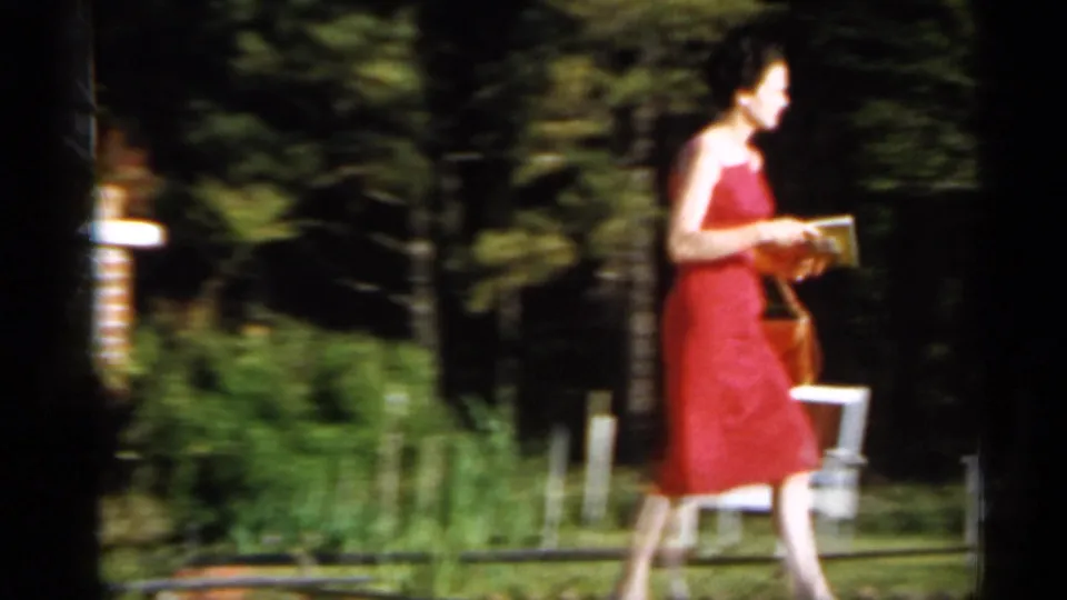 A different woman in a red dress walks through a wooded outdoor area, holding an object, in the same 1958 Florida home-movie clip.