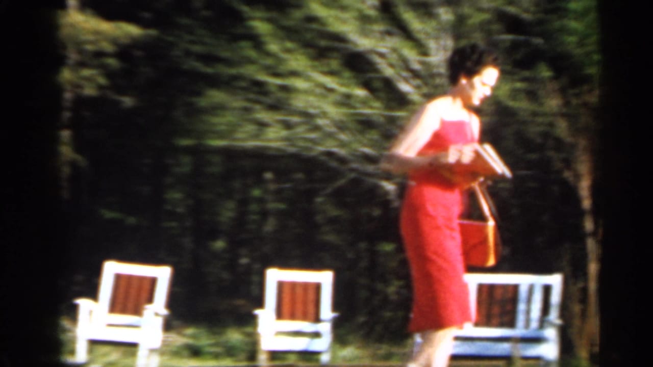 Woman in a red dress carries a book and purse near white lawn chairs in a shaded yard.