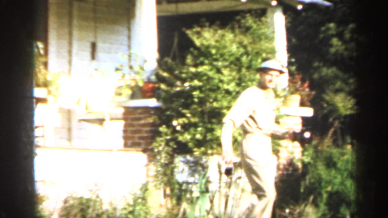 Man in a light suit and hat walks near a house exterior while holding a camera.