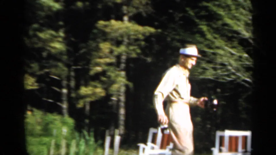 The man in a light suit and hat continues across the green yard, holding a camera with white outdoor chairs visible behind him.