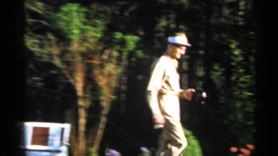 A man in midcentury dress walks through the shaded Florida yard holding a camera, with a white chair and dense greenery in the background.