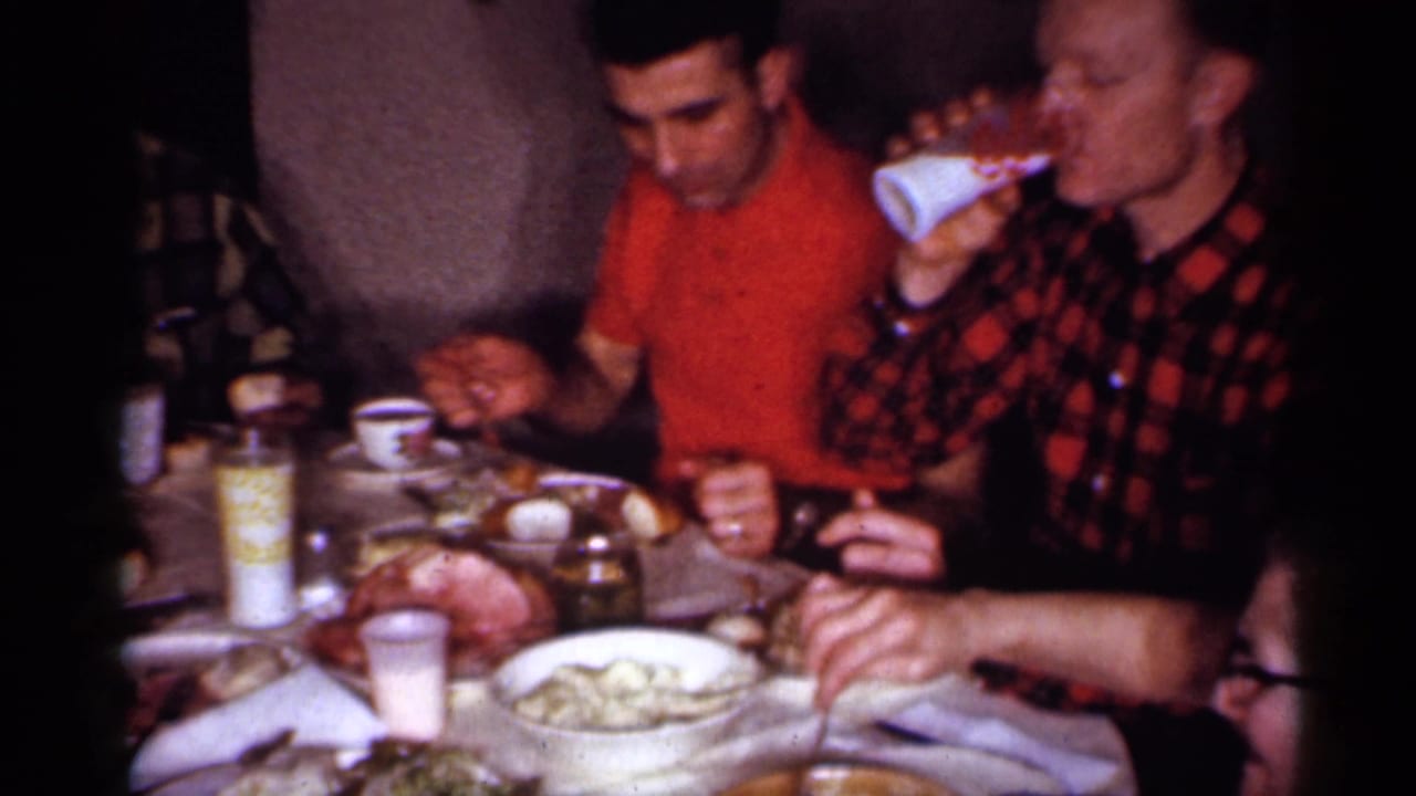1958 New Jersey home-movie frame of adults eating at an indoor dining table with food, cups, and a condiment bottle visible.