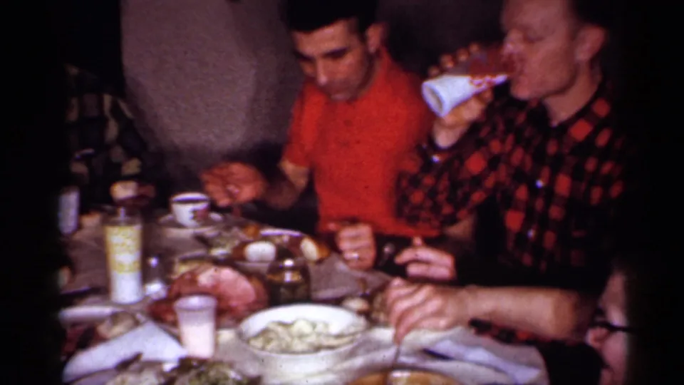1958 New Jersey home-movie frame of adults eating at an indoor dining table with food, cups, and a condiment bottle visible.