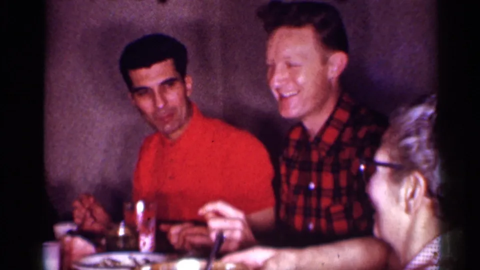 1958 New Jersey home-movie frame showing two men at a dining table and a woman in glasses in the foreground.