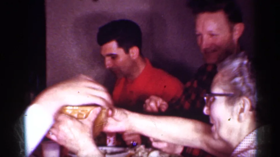 1958 New Jersey home-movie frame of a woman in glasses reaching across a dinner table with seated adults behind her.