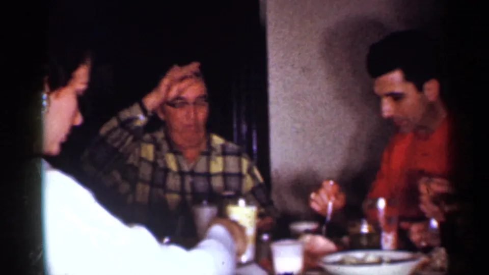 1958 New Jersey home-movie frame of adults at an indoor meal, including a man touching his forehead and tableware in front.