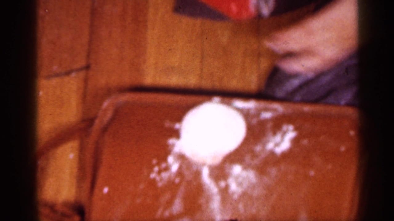1958 New Jersey home-movie frame showing white cream on a brown surface near a woman in a robe, with soft 8mm color and blur.
