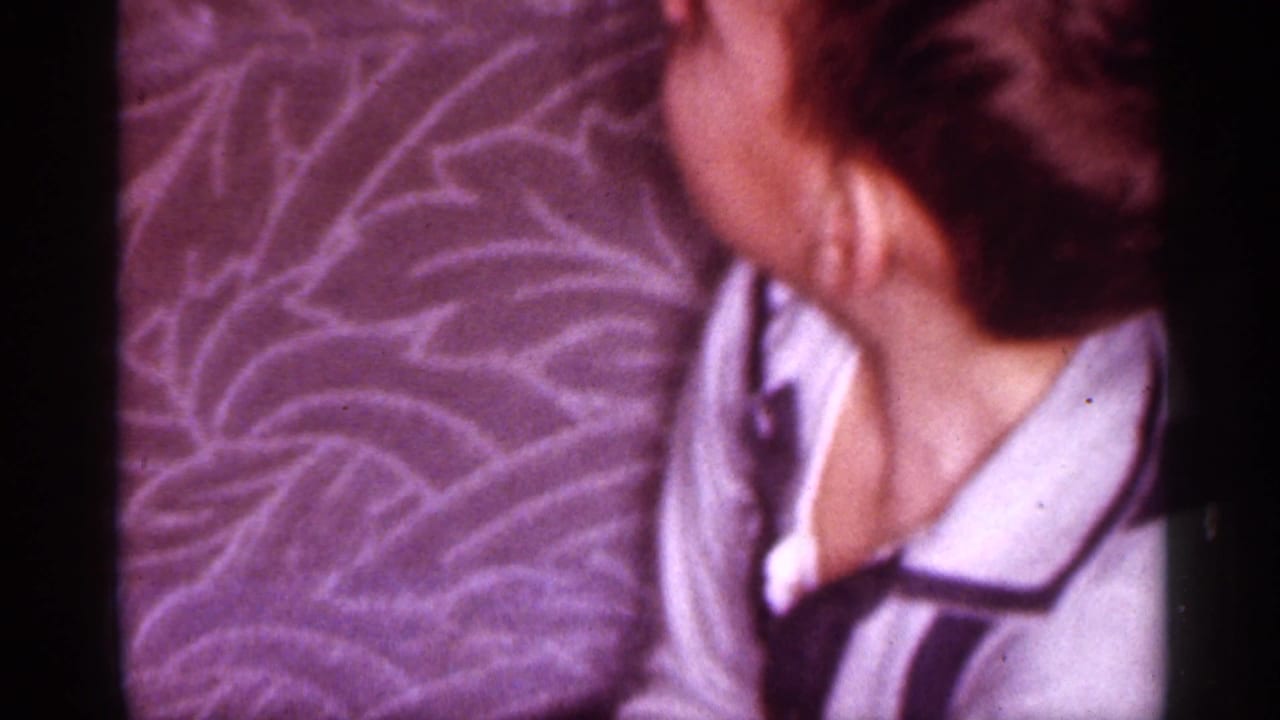 1958 New Jersey home-movie frame of a woman in a robe seated close to patterned upholstery in a private interior.
