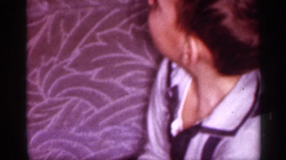 1958 New Jersey home-movie frame of a woman in a robe seated close to patterned upholstery in a private interior.