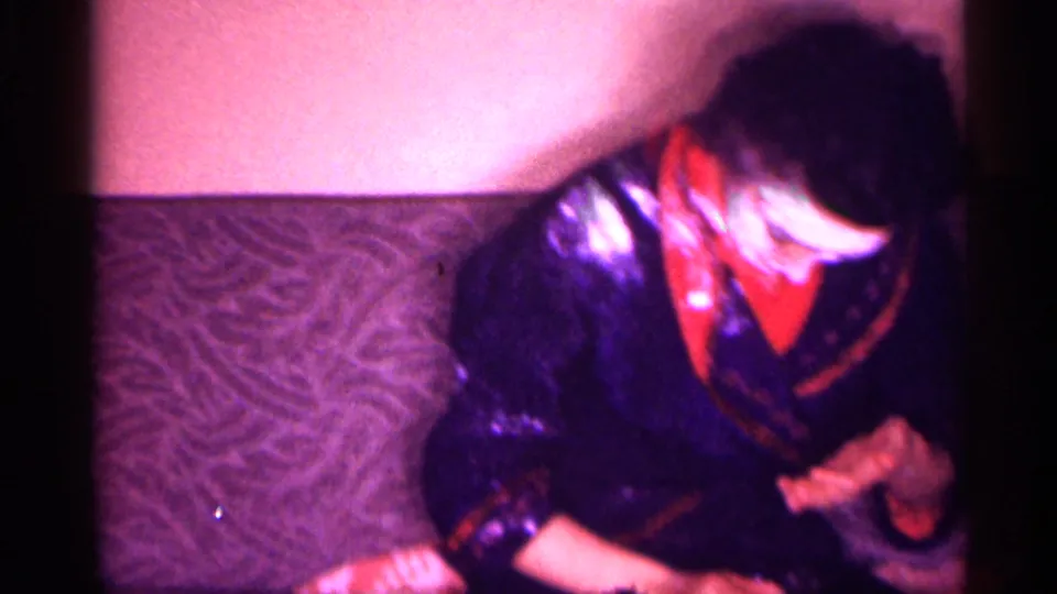 1958 New Jersey home-movie frame showing a woman in a dark blue robe with red collar applying white cream to her face.