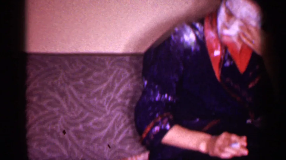 1958 New Jersey home-movie frame of a woman in a blue and red robe seated by patterned upholstery after applying cream.