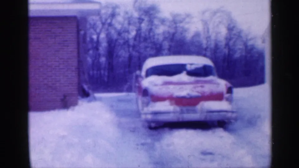 1958 New Jersey home-movie frame showing a two-tone car in a snow-covered driveway near a brick suburban house.