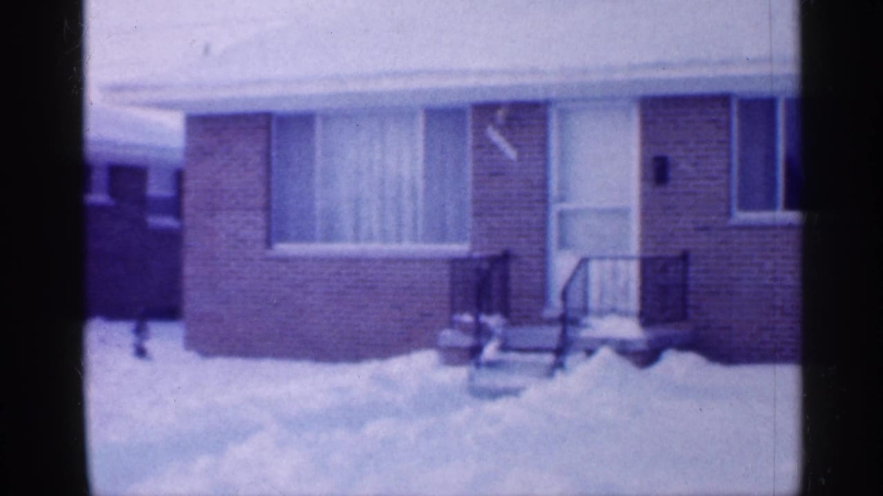 1958 New Jersey home-movie frame showing a modest brick house exterior with snow in the yard, a small porch, and curtained window.