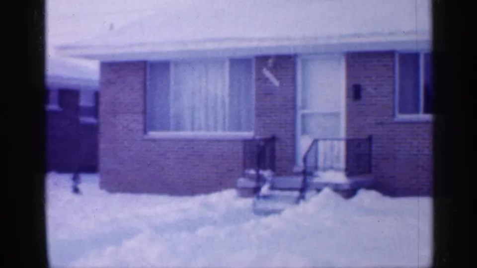 1958 New Jersey home-movie frame showing a modest brick house exterior with snow in the yard, a small porch, and curtained window.
