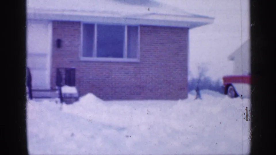 1958 New Jersey winter frame showing a brick house, snow-covered yard, bare trees, and part of a red vehicle at the edge.