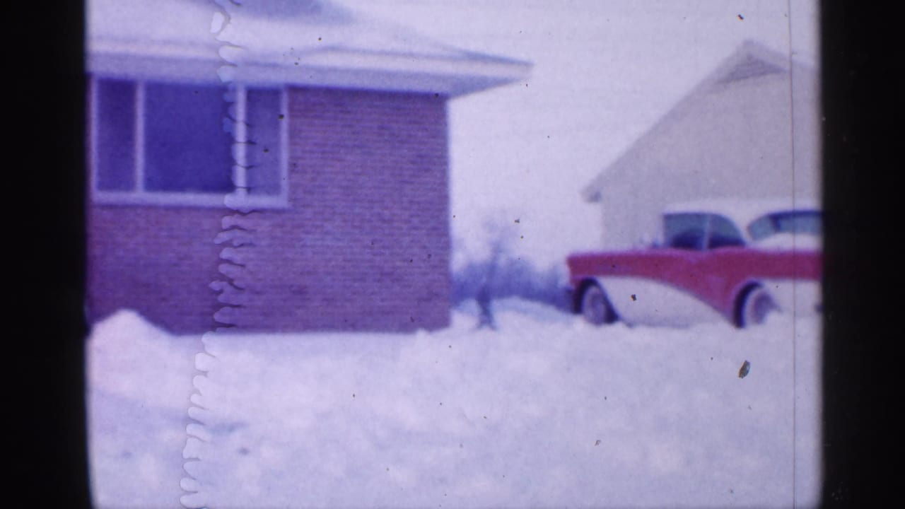 Snowy driveway beside a brick house with a red-and-white car partly buried in snow.
