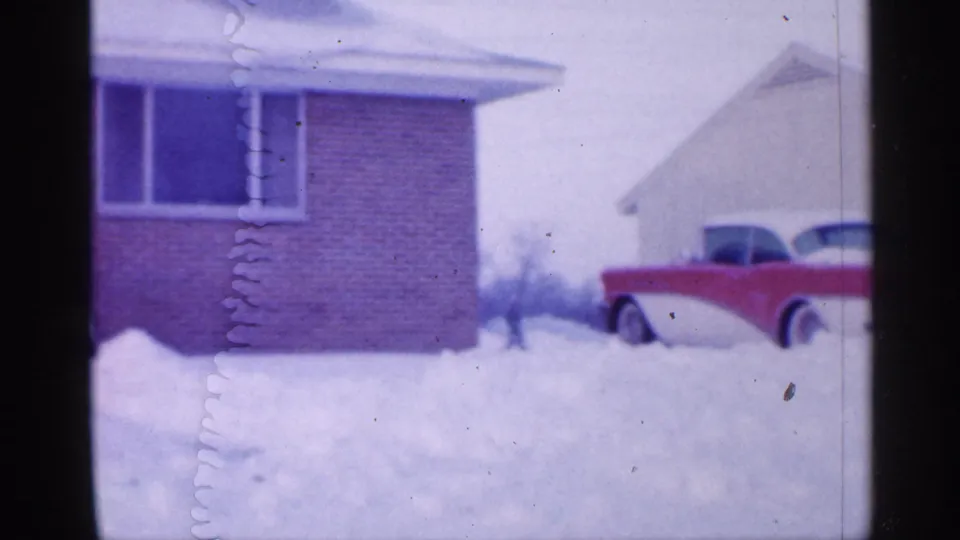 1958 New Jersey home-movie frame showing a red-and-white 1950s car in deep snow near a brick house and neighboring white house.