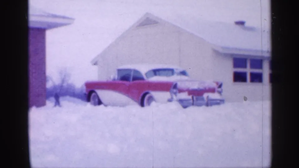 The snowbound red-and-white car is seen more clearly near a white neighboring house, with heavy snow covering the driveway area.