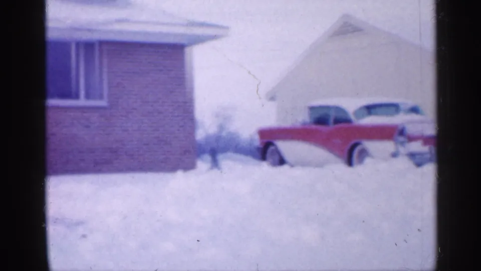 A soft 8mm view shows the brick house at left and the red-and-white car partly cropped by the frame amid deep winter snow.