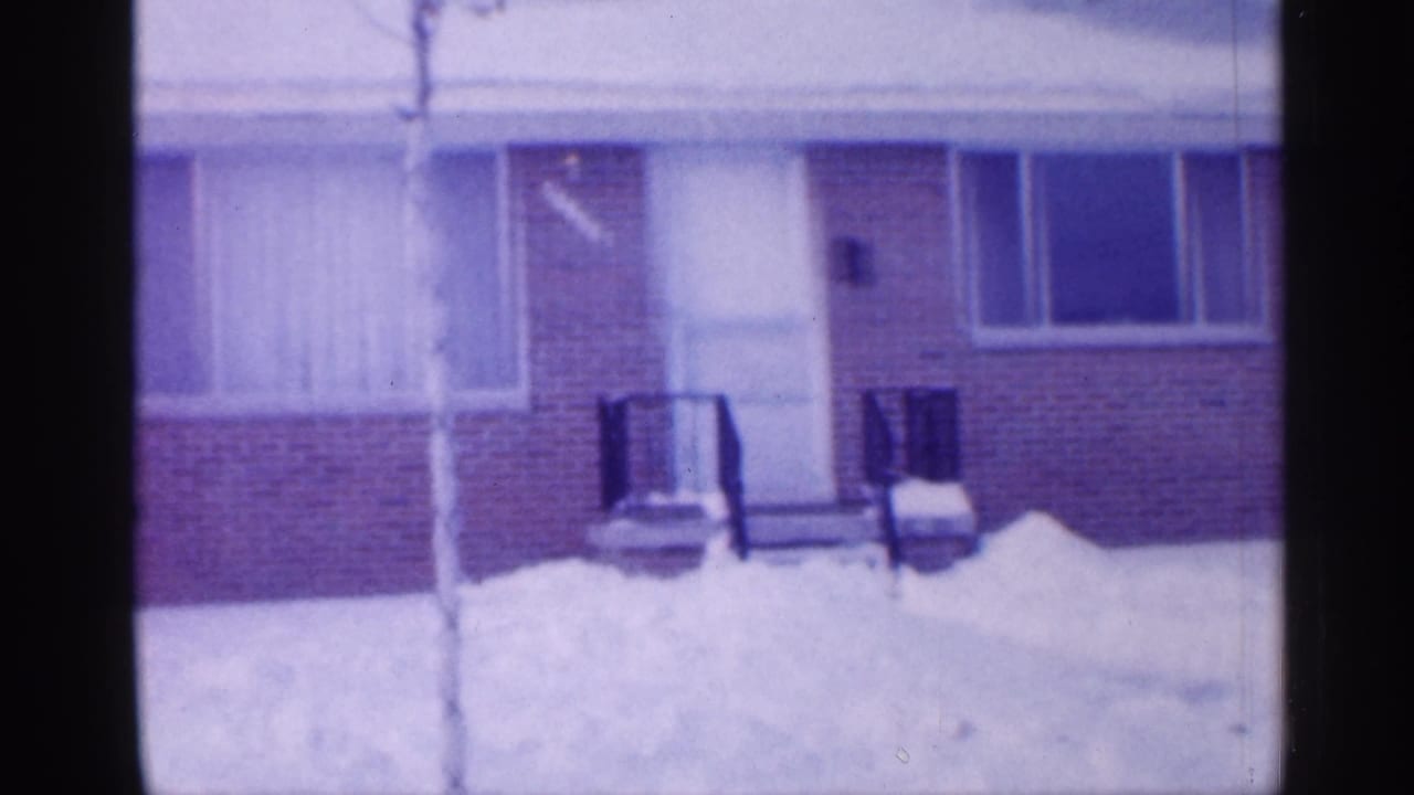 1958 New Jersey home-movie frame showing a brick suburban house front behind deep snow and bare foreground branches.