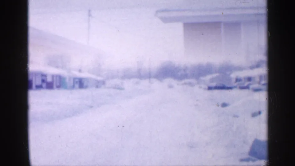 Soft 8mm frame of a snow-covered New Jersey suburban street in 1958, with low homes and parked cars partly obscured by exposure haze.