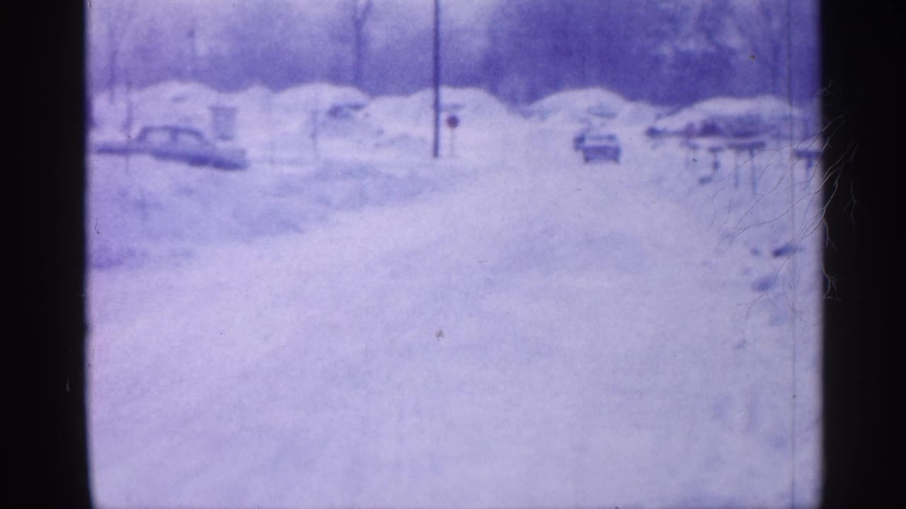 1958 New Jersey home-movie frame of a snowy street with parked mid-century cars, bare trees, and utility poles in soft blue-purple 8mm color.