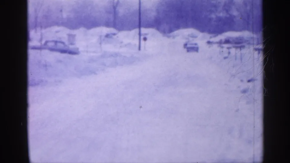 1958 New Jersey home-movie frame of a snowy street with parked mid-century cars, bare trees, and utility poles in soft blue-purple 8mm color.