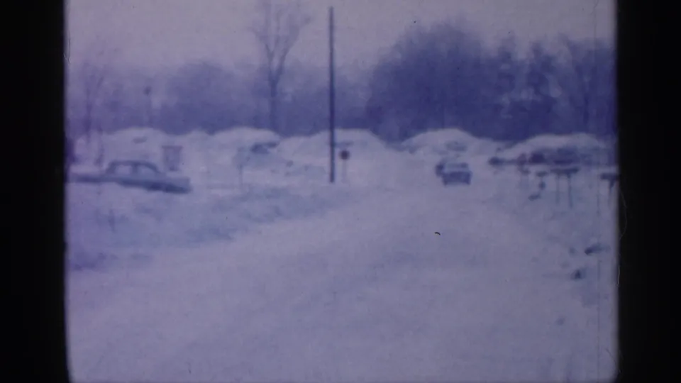 A soft 8mm view of a snowy New Jersey residential street in 1958, with cars partly surrounded by snowbanks and houses in the background.