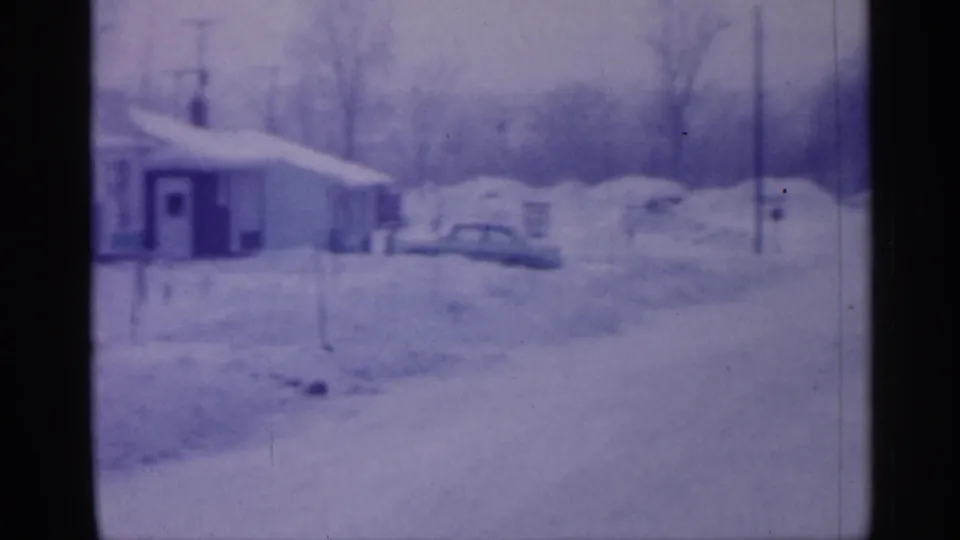 A modest housefront and parked cars appear along a snowy New Jersey street, recorded in blue-purple 8mm home-movie color in 1958.