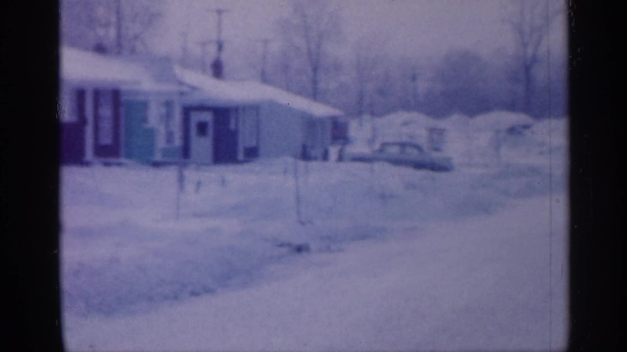 Snow-covered residential street with houses, parked cars, bare trees, and deep snowbanks.