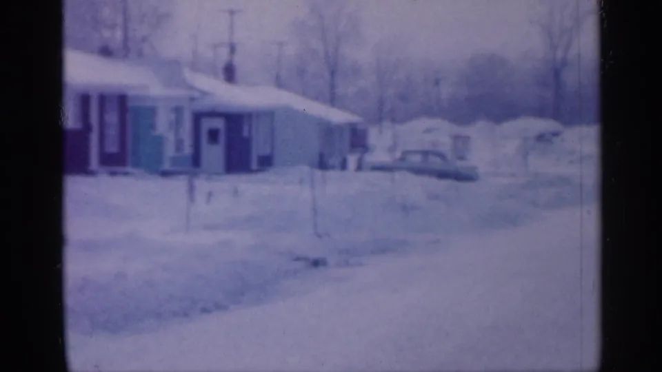 1958 New Jersey home-movie frame showing a snowy residential block with low houses, buried cars, bare trees, and a blue-purple film cast.