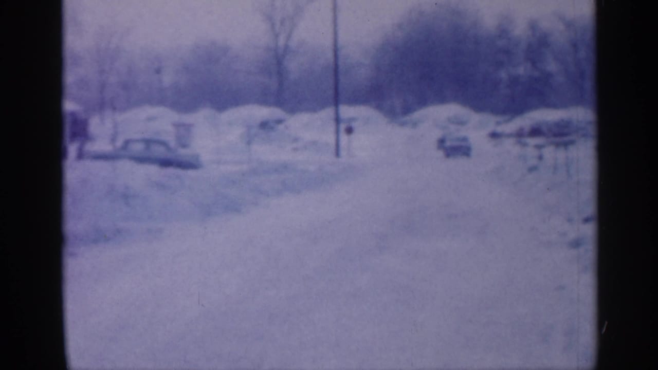 Snowy street view with buried cars, distant houses, utility poles, and bare trees.