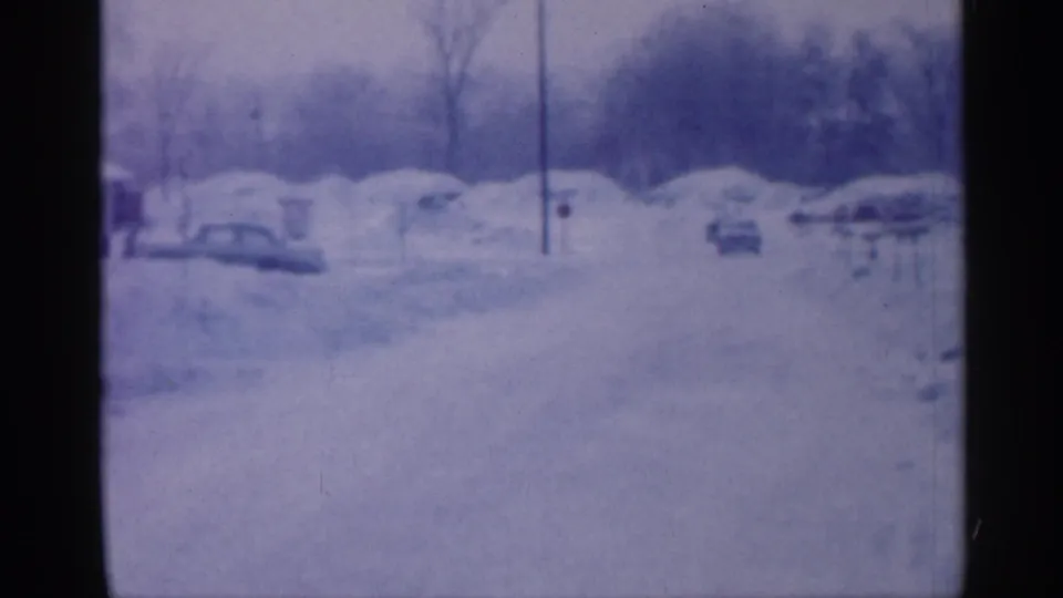 A 1958 New Jersey winter street appears under heavy snow, with parked cars, low houses, poles, and bare trees softened by 8mm grain.