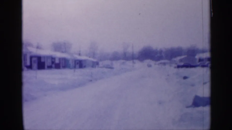 Wide 8mm frame of a New Jersey residential neighborhood in 1958, with houses, snow-covered road, parked cars, bare trees, and heavy drifts.