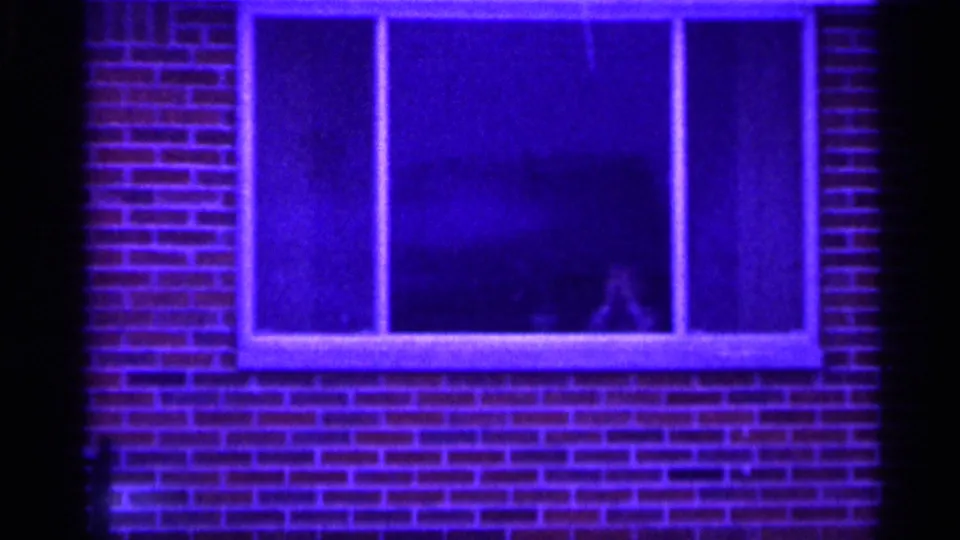 Blue-tinted 1958 home-movie frame of a brick facade and wide window with a shadowy person partly visible inside.