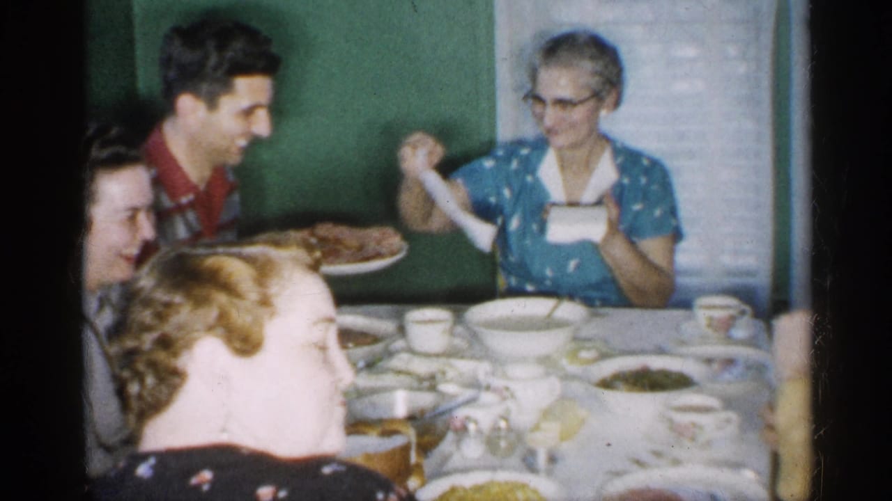 In a 1958 New Jersey home-movie frame, family members sit around a dining table while a man in a red shirt holds a platter of food and an elderly woman in a blue dress handles a napkin.