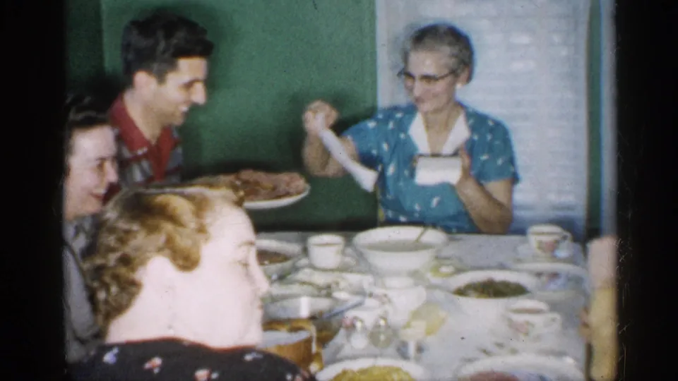 In a 1958 New Jersey home-movie frame, family members sit around a dining table while a man in a red shirt holds a platter of food and an elderly woman in a blue dress handles a napkin.