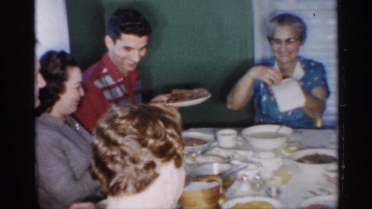 The man in a red patterned shirt holds a platter near seated relatives as the elderly woman in a blue dress extends a napkin across the dinner table.