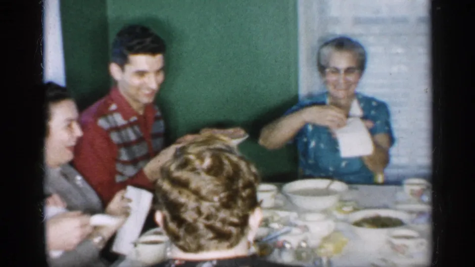 At the dinner table, the man holding a platter smiles toward seated relatives while the elderly woman in blue holds a napkin beside bowls, cups, and dishes.