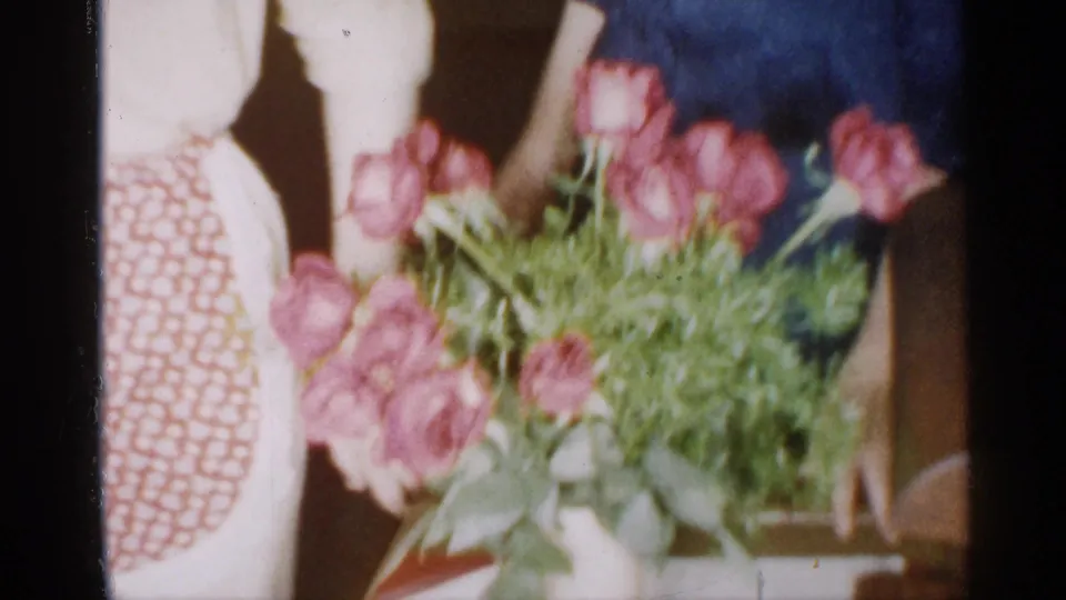 Pink flowers sit in the foreground of a 1958 New Jersey home-movie frame, with a red patterned apron and table area visible behind them.