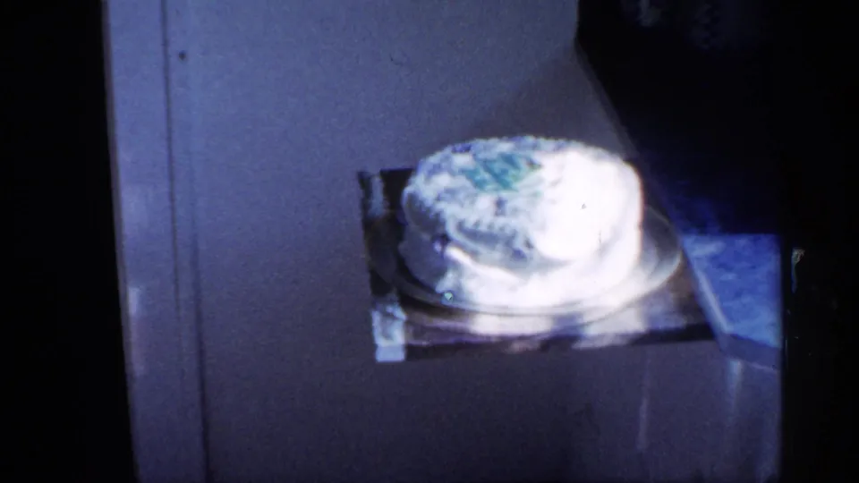 A white frosted cake sits on a counter or table in a 1958 New Jersey home interior, with a person partly visible at the edge.
