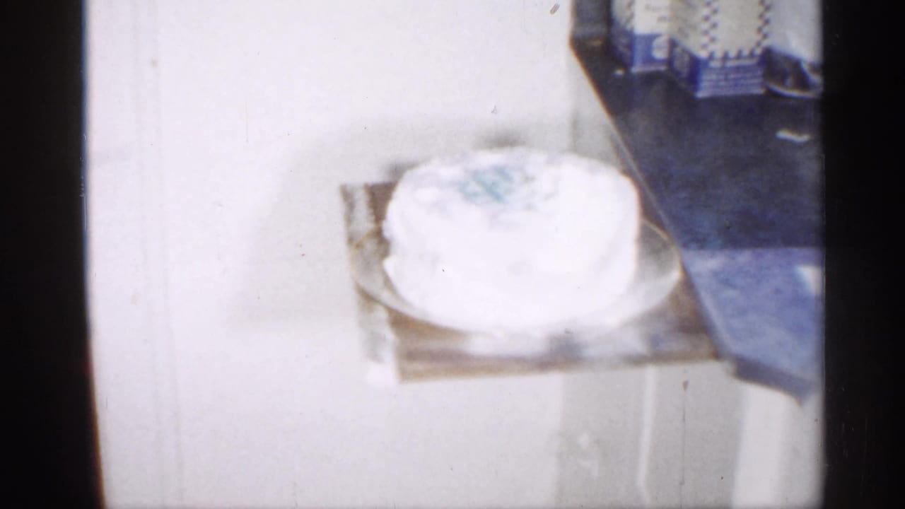 A white frosted cake with pale blue decoration rests on a table in a 1958 New Jersey home-movie interior.