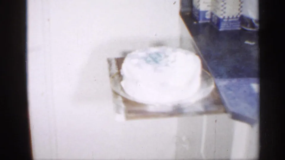A white frosted cake with pale blue decoration rests on a table in a 1958 New Jersey home-movie interior.
