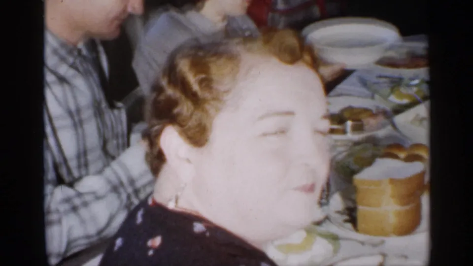 A close, overexposed home-movie view shows a smiling woman and other adults beside plates, cups, and food at the dinner table.